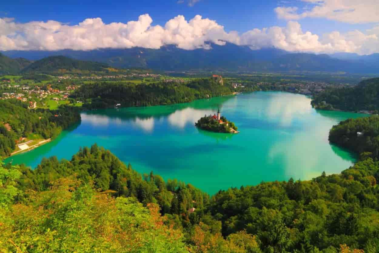 Lake Bled island church surrounded by turquoise water, green hills, and mountains under a blue sky.