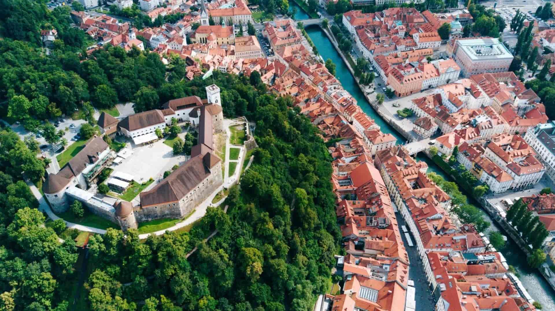 Aerial view of Ljubljana Castle above dense green trees and the city's red-roofed buildings and river.