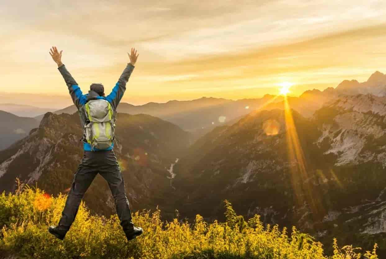 Hiker with backpack jumping on mountain ridge at sunset over valley