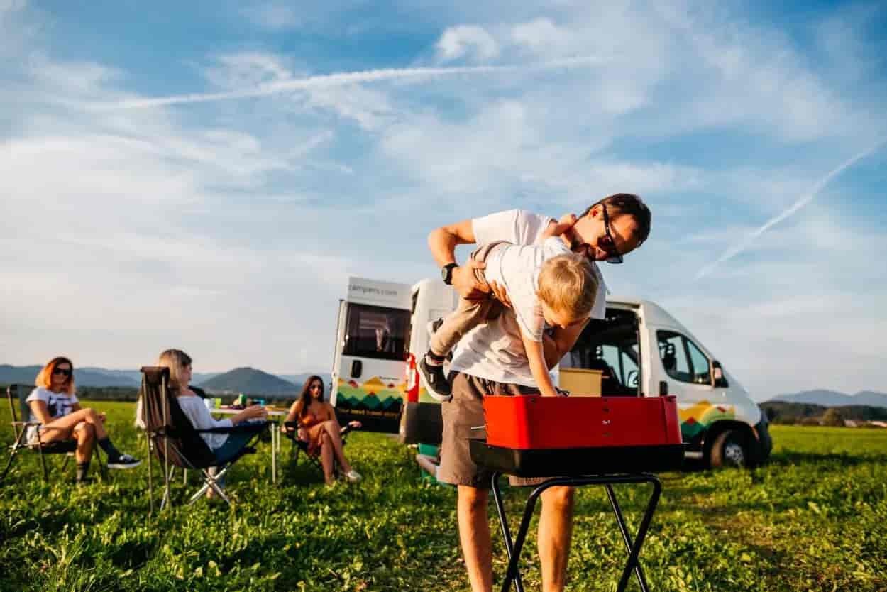 Family camping with camper van, grilling outdoors in grassy field with mountains in background.