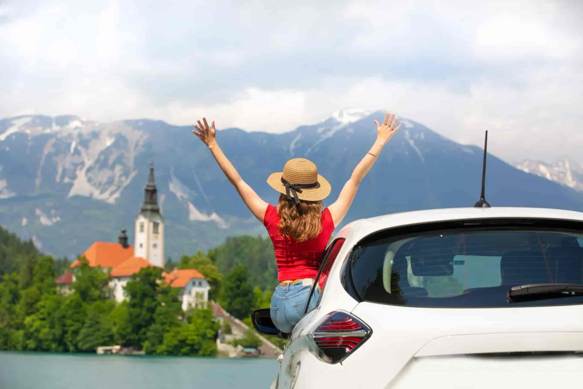 Woman with arms raised leans out of a car window overlooking Lake Bled and mountains.