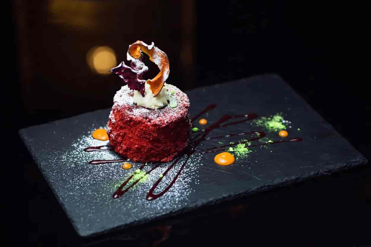 Red velvet cake with cream topping, powdered sugar, and sauces on a dark slate platter