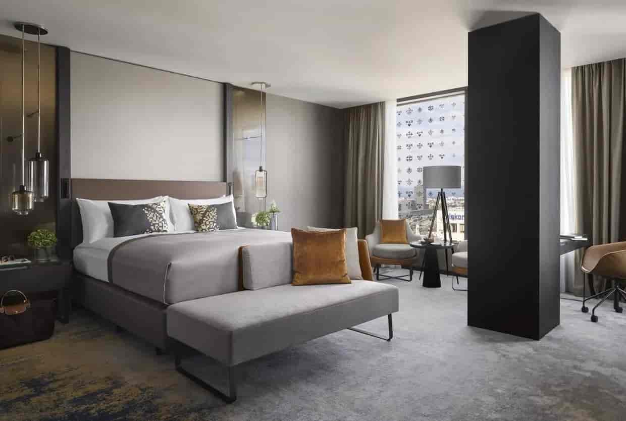 Modern hotel king deluxe room with gray bedding, accent pillows, and city view through window.