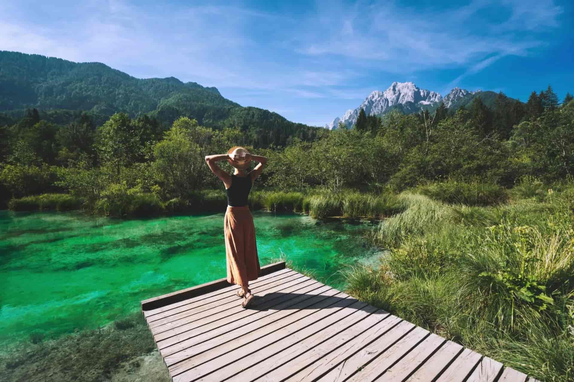 Woman with hat on wooden dock overlooking emerald green spring water and mountains