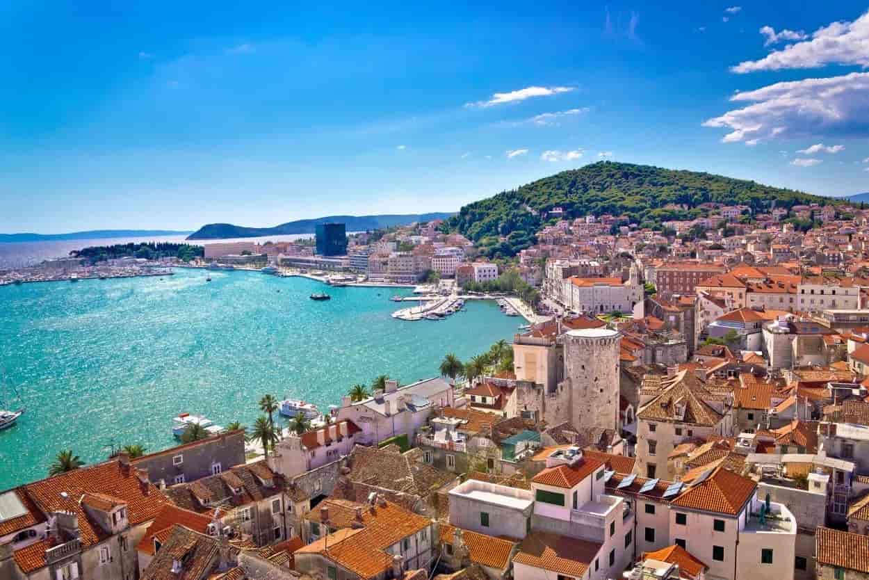 Coastal city with terracotta roofs and harbor on bright blue water under a sunny sky