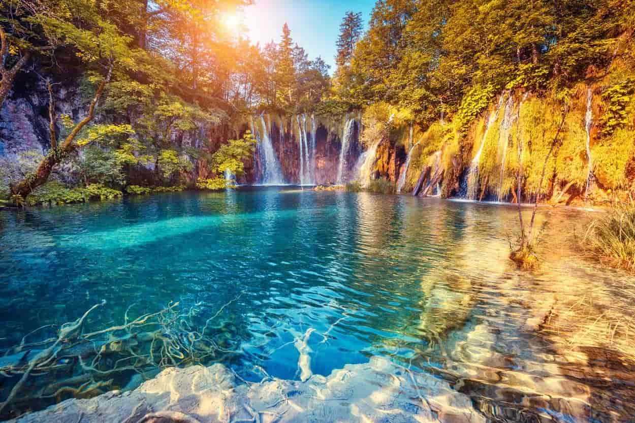 Plitvice Lakes waterfalls cascading into turquoise pool with sunlit autumn foliage.