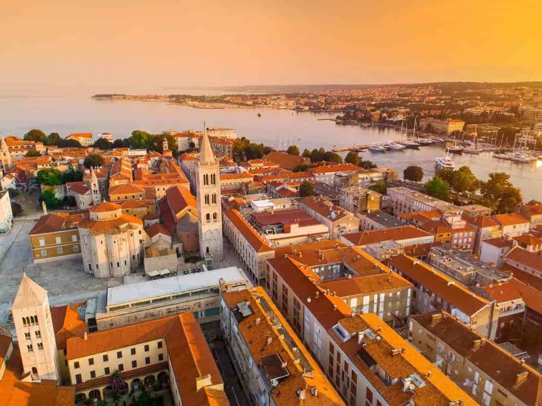 Aerial view of Zadar old town with terracotta roofs, bell tower, and harbor at sunset.