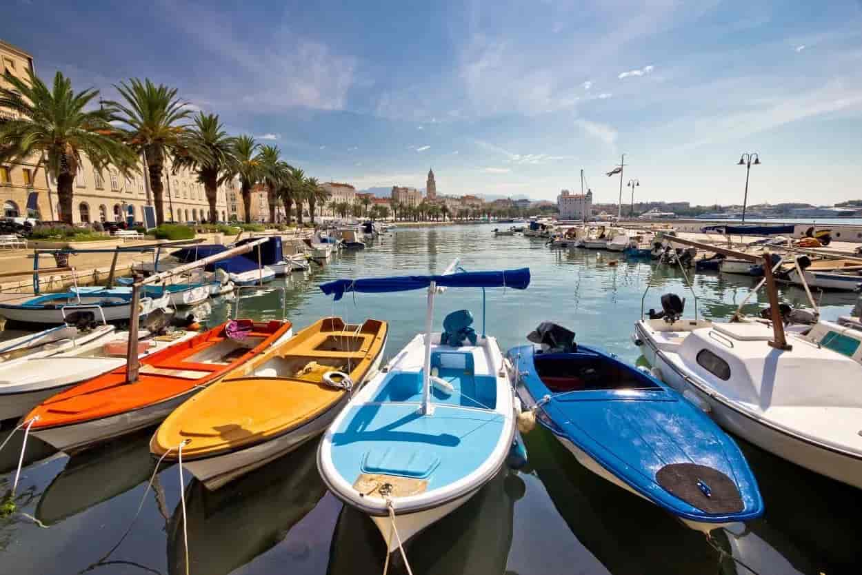 Colorful small boats moored in a harbor with palm trees and historic buildings in the background.