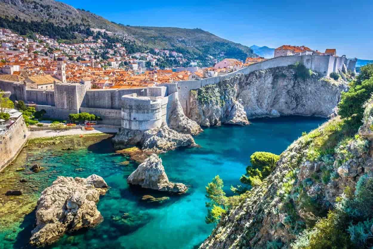 Dubrovnik city walls overlooking turquoise Adriatic Sea cove and hillside town.