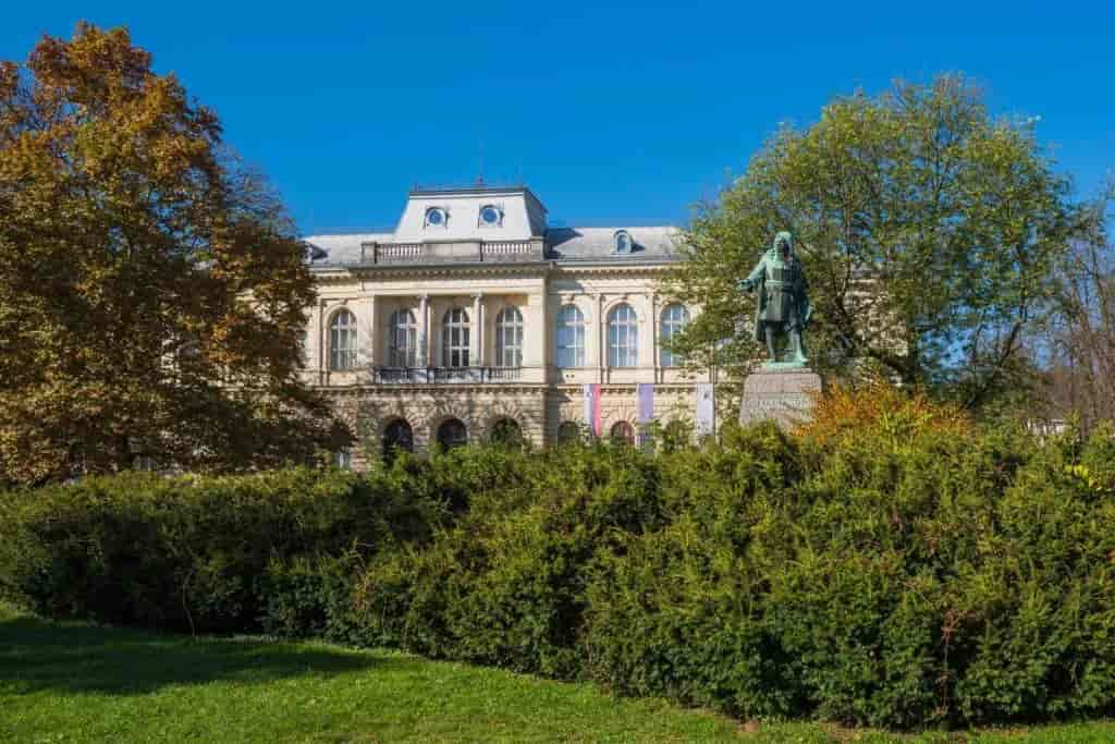 Ljubljana National Museum building with statue in foreground garden under clear blue sky.