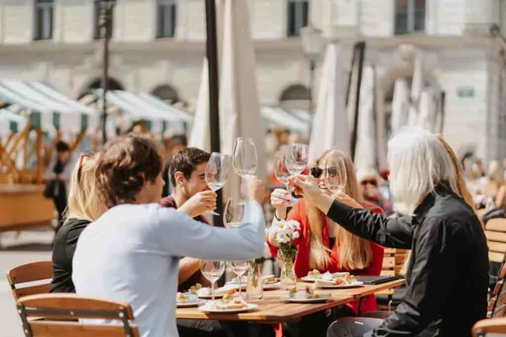Group toasting with wine glasses at an outdoor cafe table with food in a sunny city square.