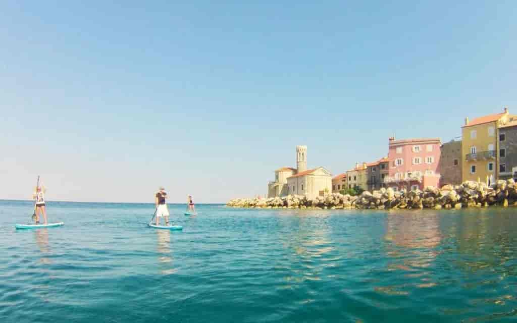 Stand-up paddleboarding near colorful coastal buildings and a stone church in Piran, Slovenia.