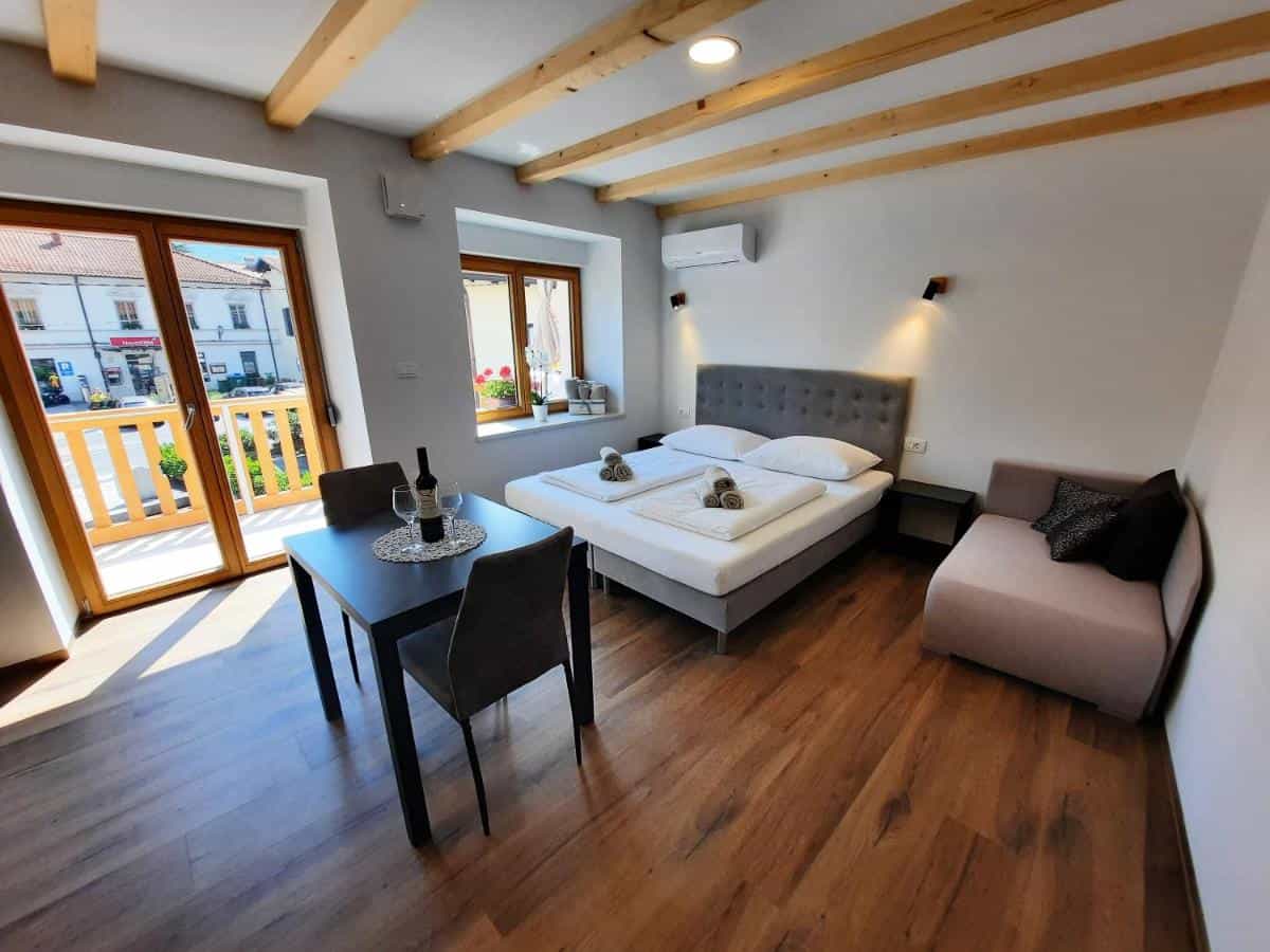 Studio apartment interior with wooden beams, double bed, dining table, and balcony view of town.