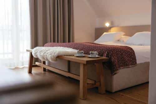 Cozy hotel bedroom with wooden bench, white sheepskin rug, and coffee cups by the bed.