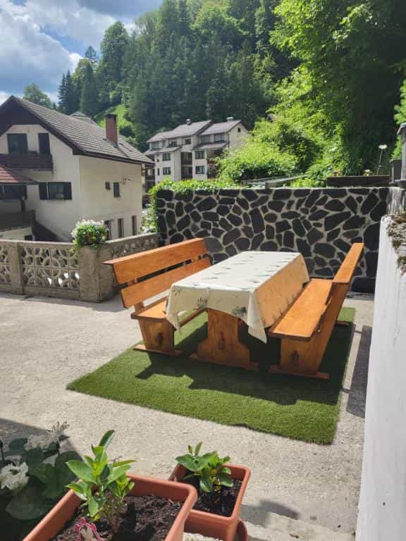 Outdoor dining area with wooden table and benches on turf, houses and forest hillside background