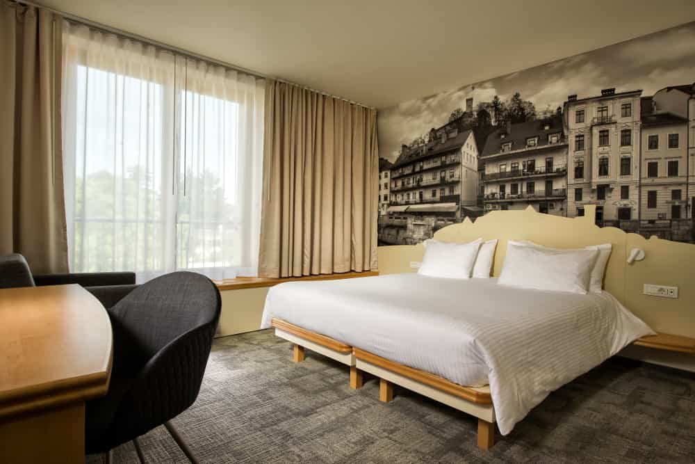 Hotel room with white bedding, desk, and black and white city mural, likely Ljubljana.