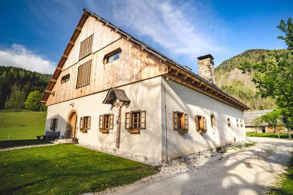 Traditional farmhouse with stucco walls and wood gable set against green mountains under blue sky.