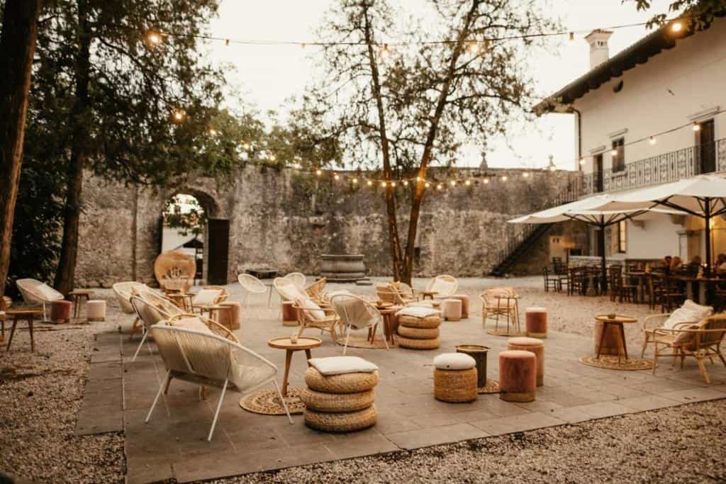 Outdoor lounge seating area with string lights against a stone wall near a building.