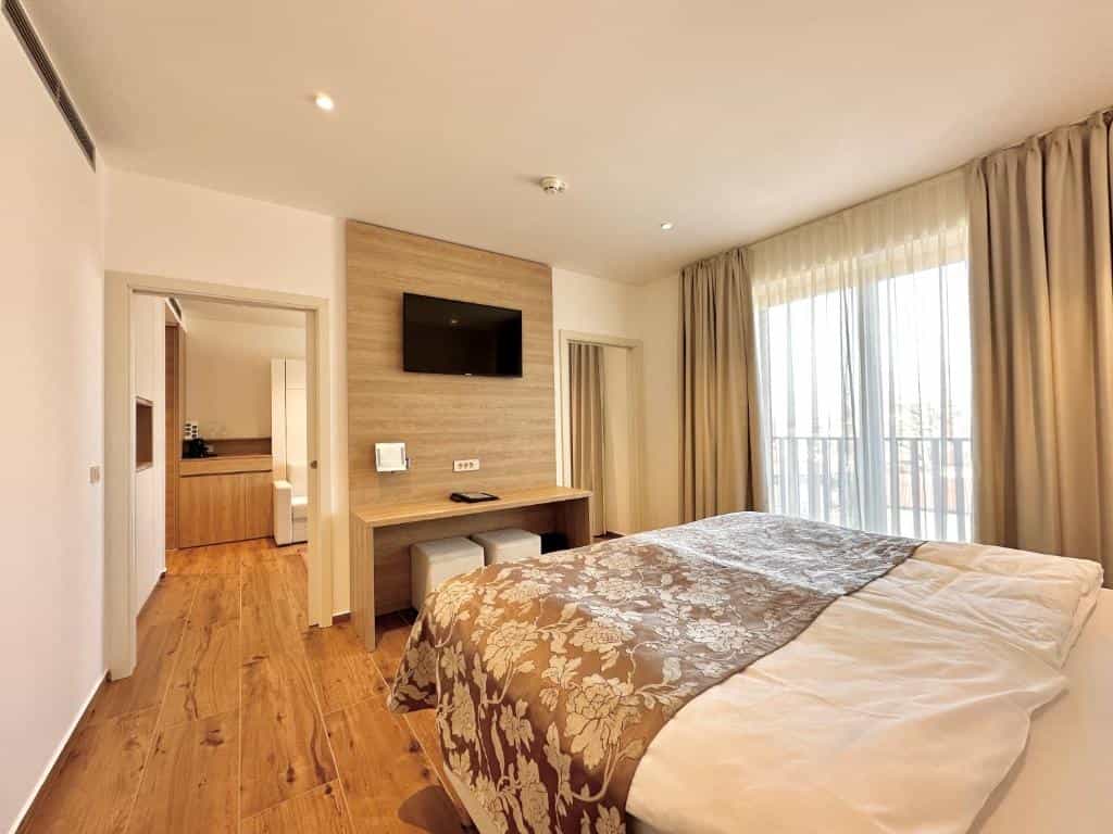 Modern hotel bedroom with wood floors, wall-mounted TV, and balcony access through sheer curtains.