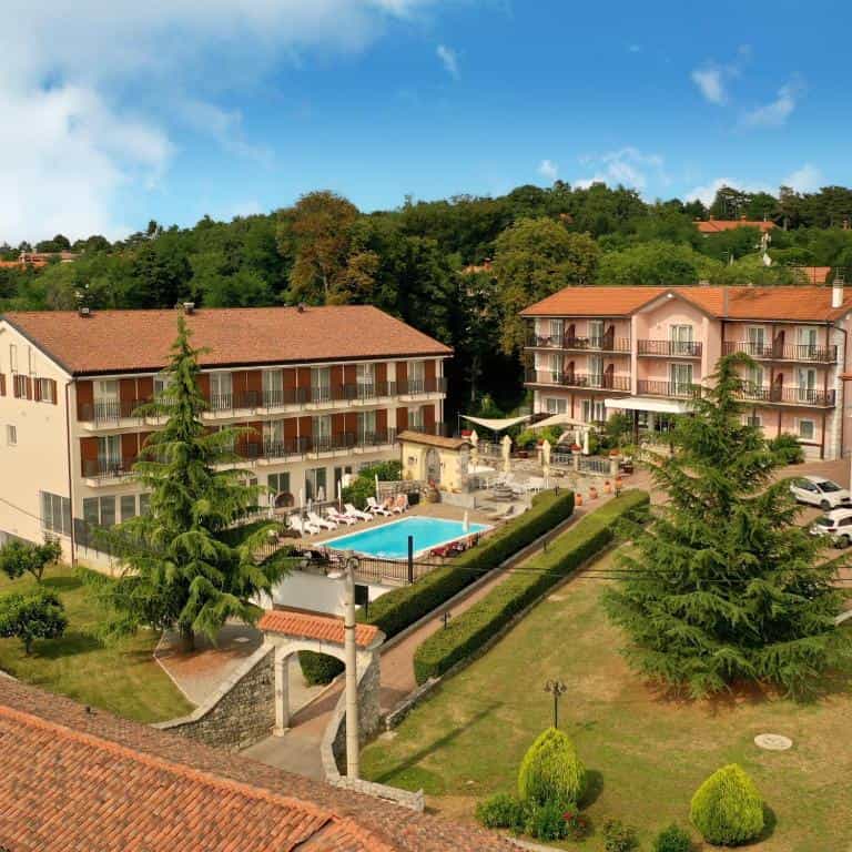 Hotel complex with two buildings, outdoor swimming pool, and green hillside setting under blue sky.