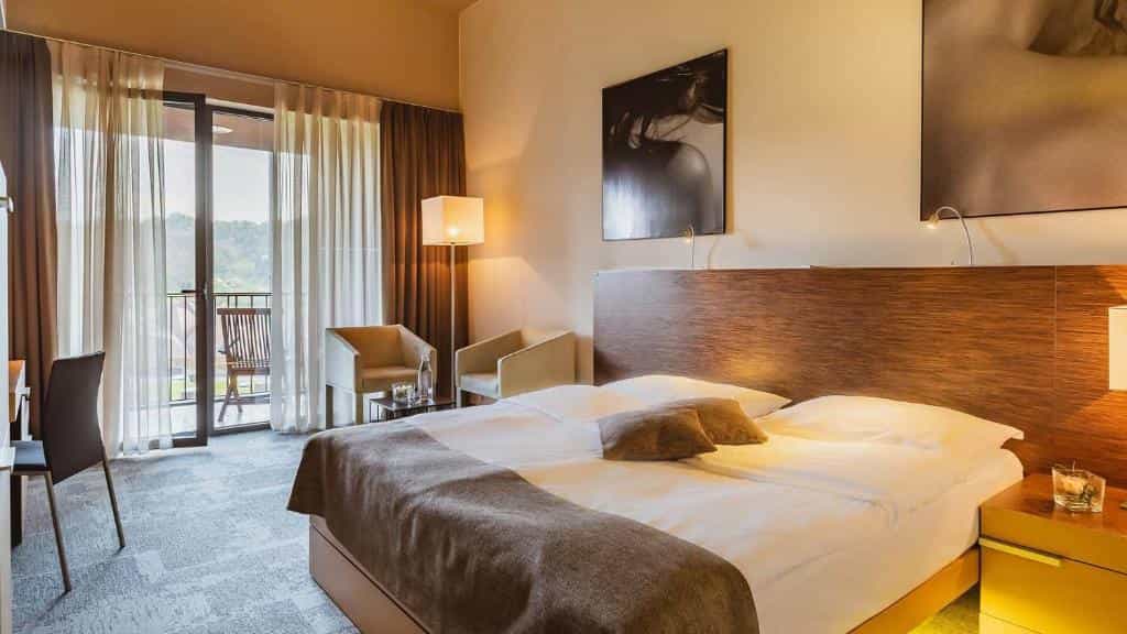 Hotel bedroom with large bed, modern decor, and balcony view of green hills.