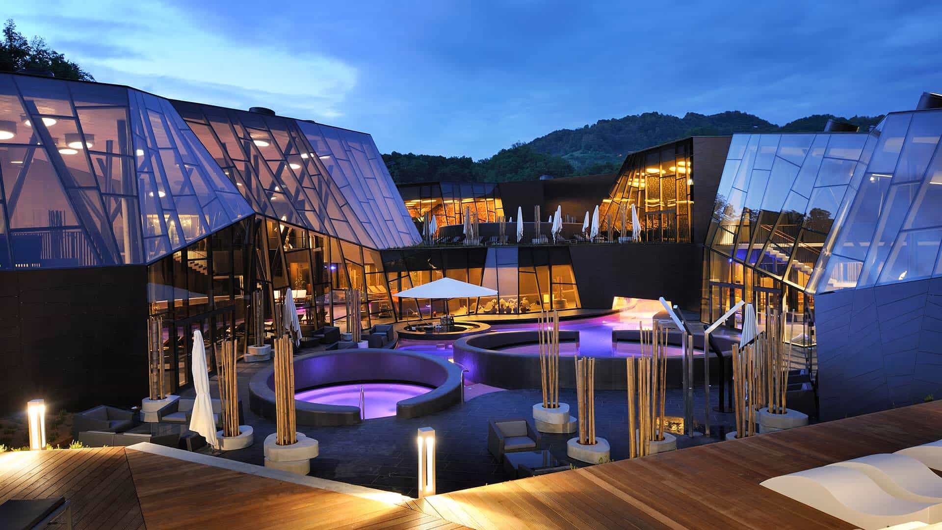Modern spa complex with geometric glass architecture and illuminated purple outdoor pools at dusk.