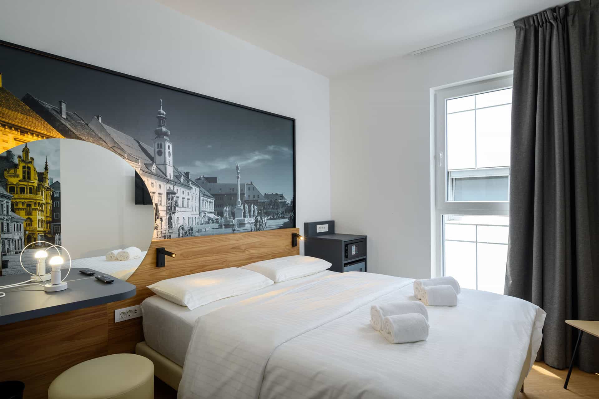 B&B Hotel Maribor room with white bedding, city mural headboard, and window with dark curtains.
