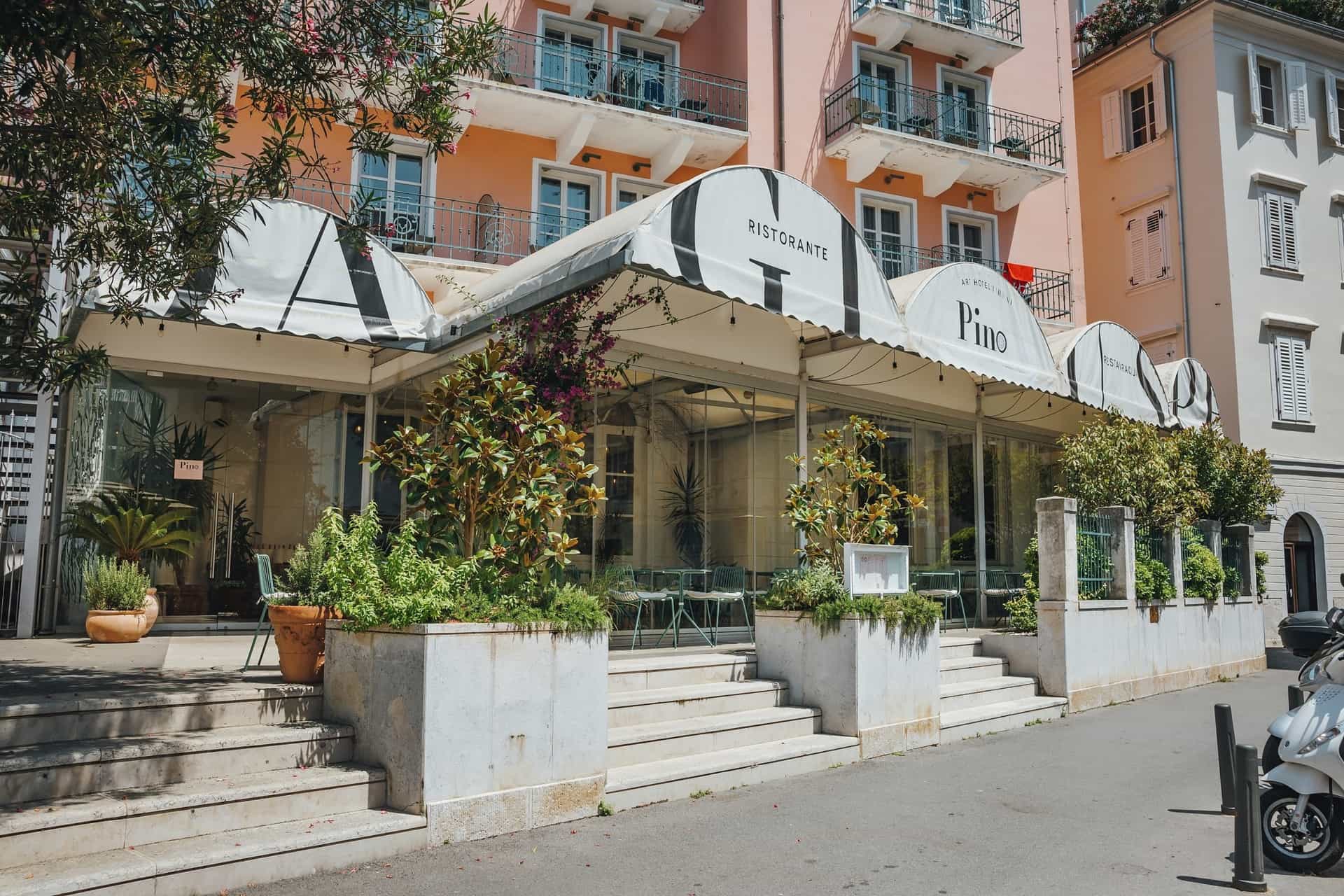 Ristorante Pino entrance with striped awning, potted plants, and pink hotel facade in background.