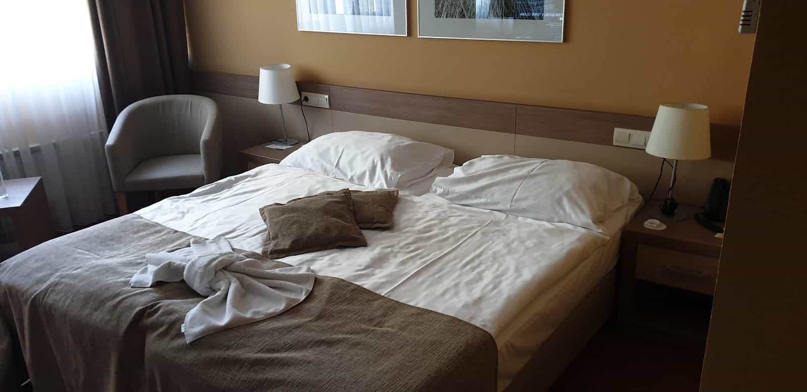 Hotel bedroom with made bed, white linens, brown throw, and bedside lamps near window.