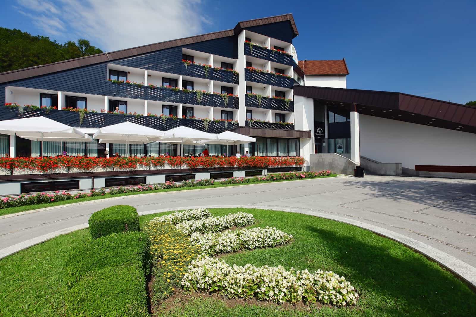 Hotel Breza exterior with balconies, red flowers, white umbrellas, and manicured lawn.