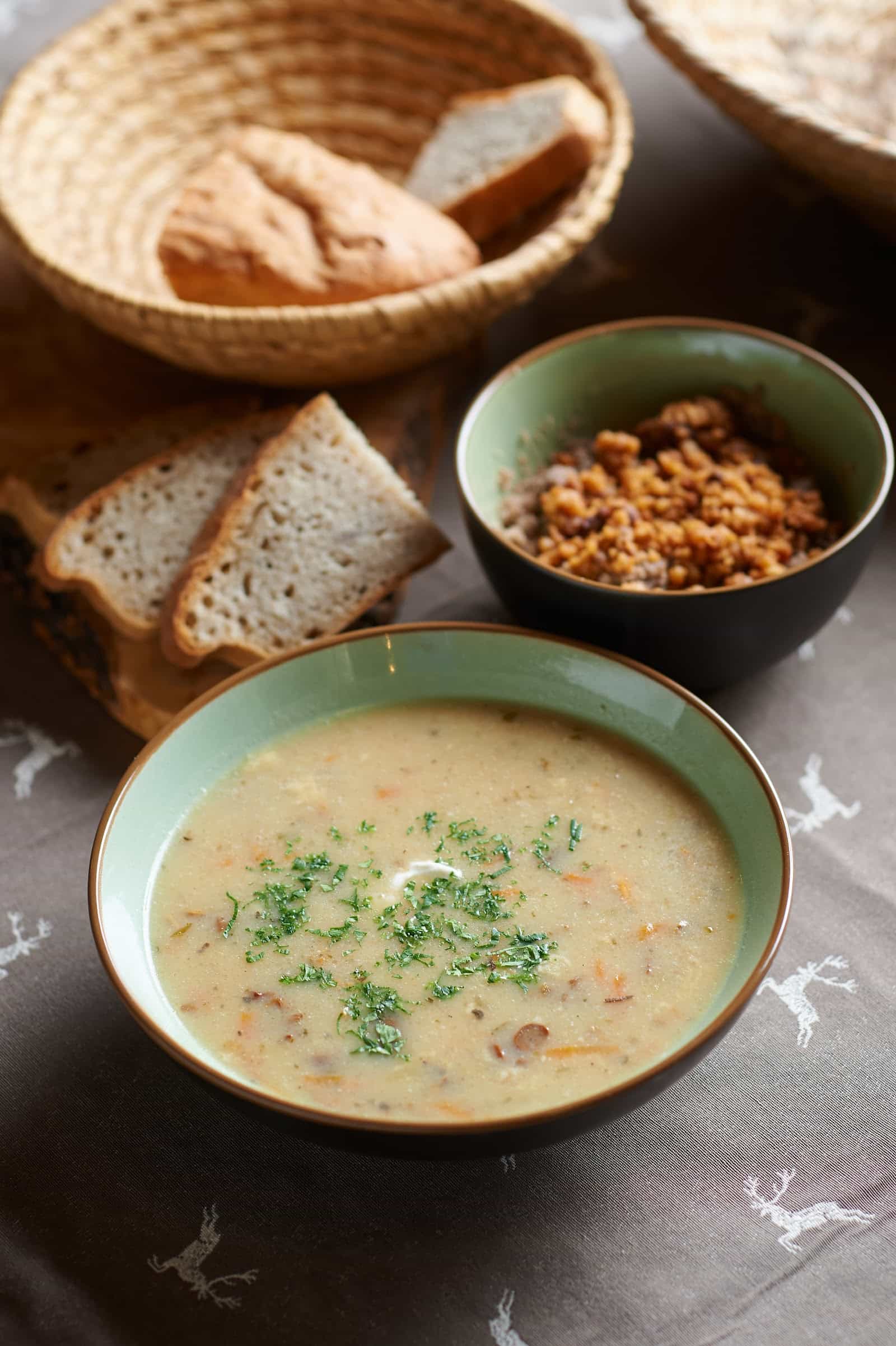 Creamy vegetable soup in a bowl with bread and a side dish on a patterned tablecloth.
