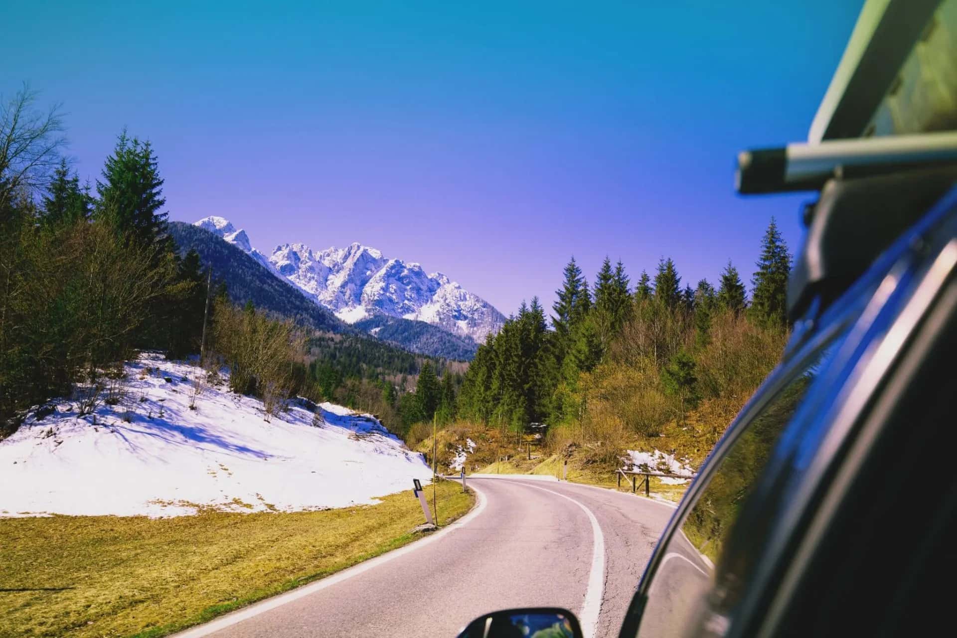 Driving on a winding mountain road past snow patches toward jagged, snow-capped peaks.