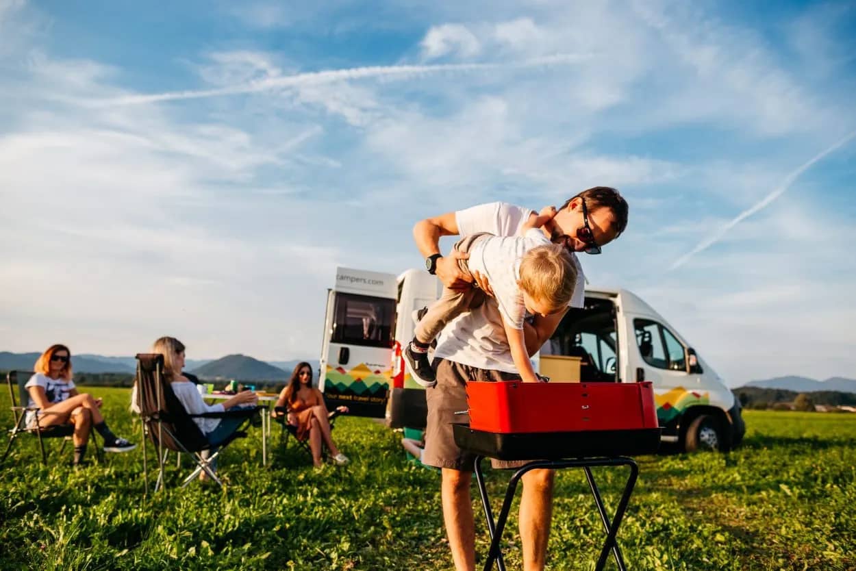 Family camping with camper van, grilling outdoors in grassy field with mountains in background.