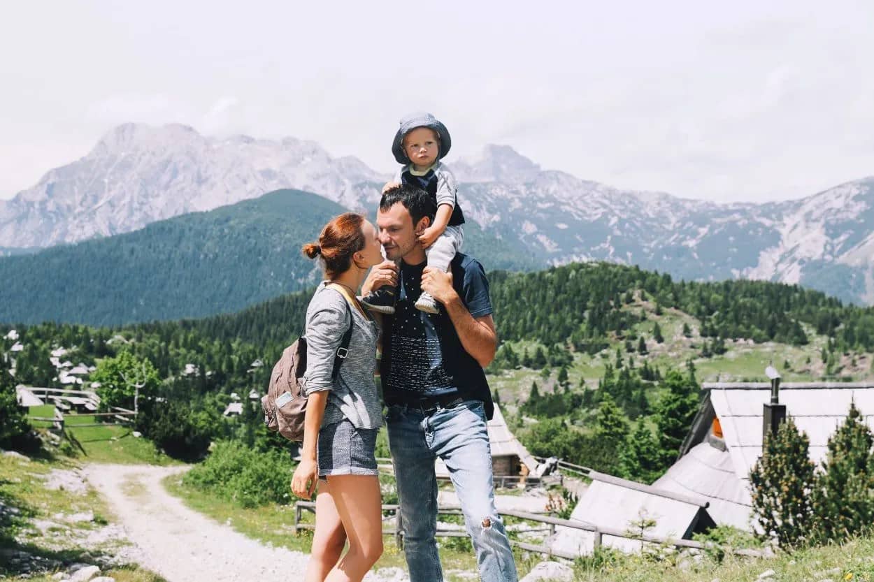 Family hiking with child on shoulders against backdrop of rocky mountains and green forest.