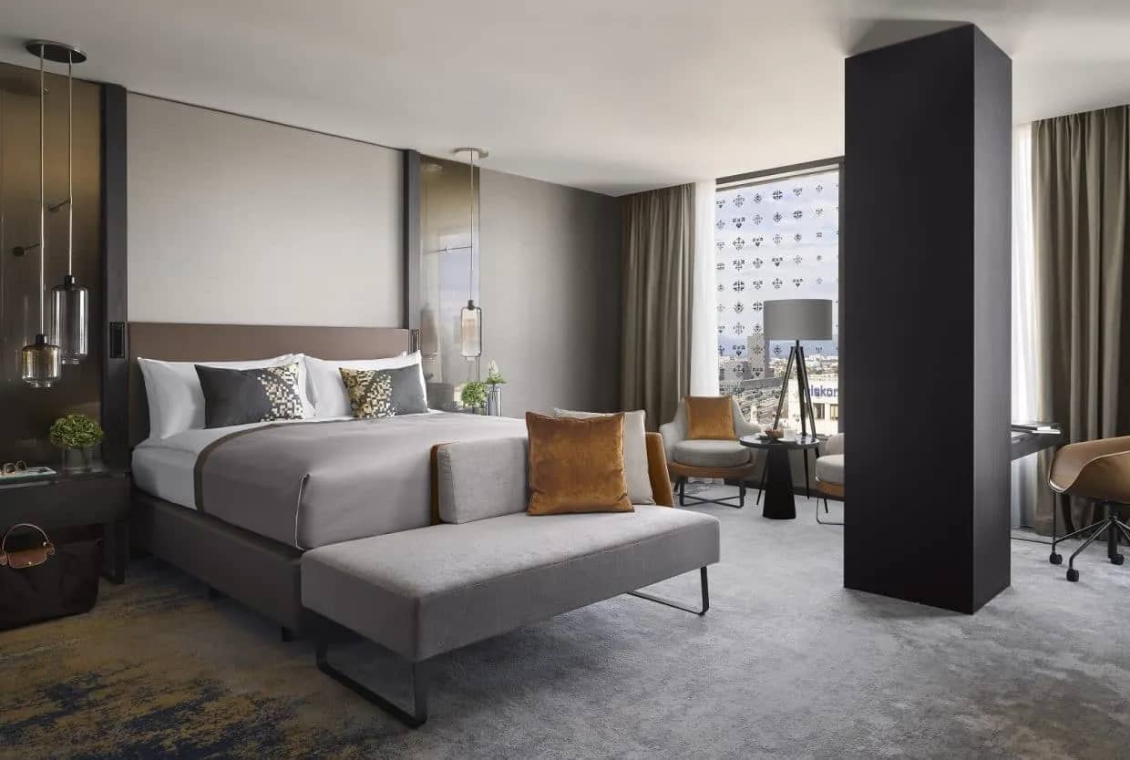 Modern hotel king deluxe room with gray bedding, accent pillows, and city view through window.