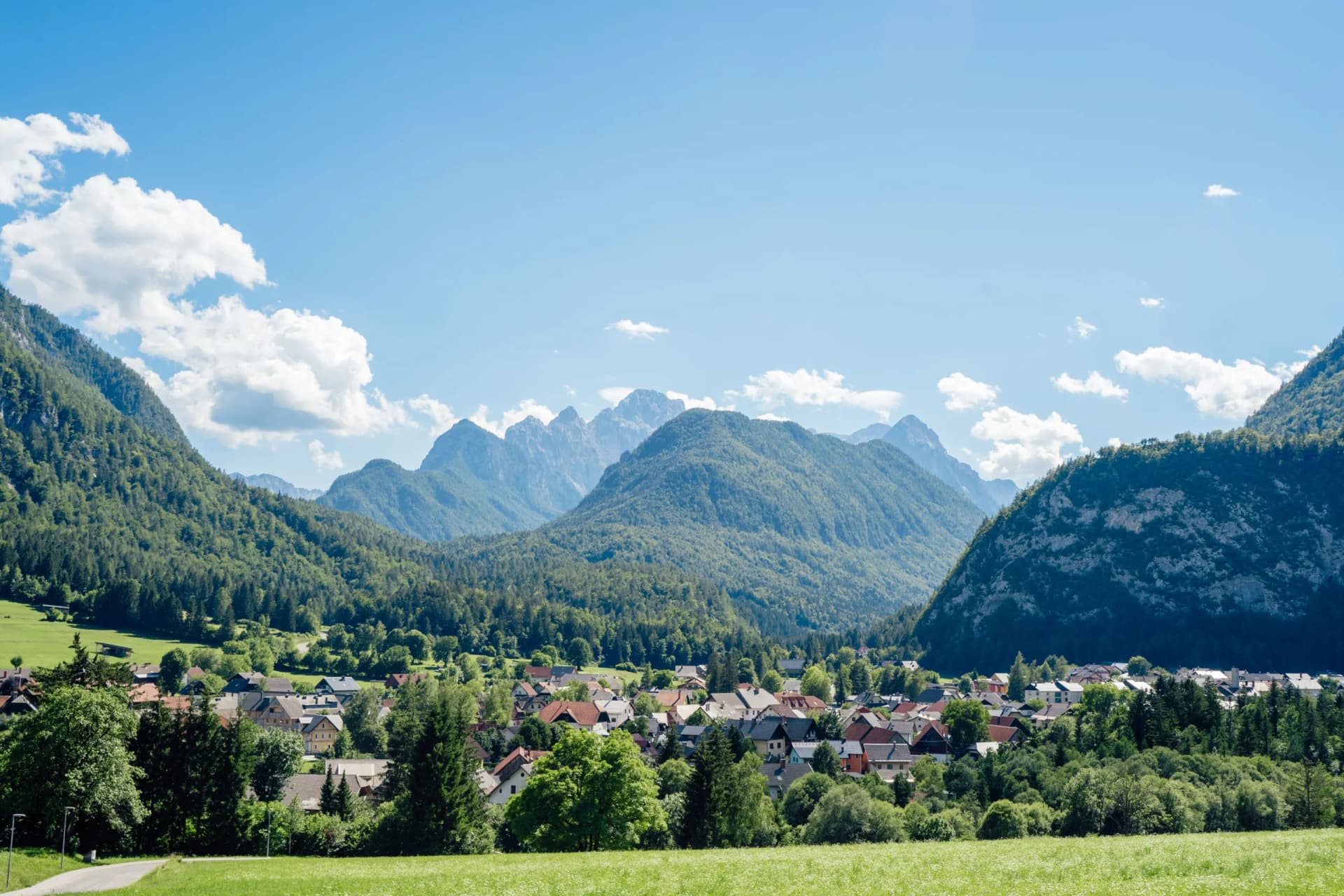 Alpine village nestled in a valley surrounded by lush green, forested mountains under a blue sky.