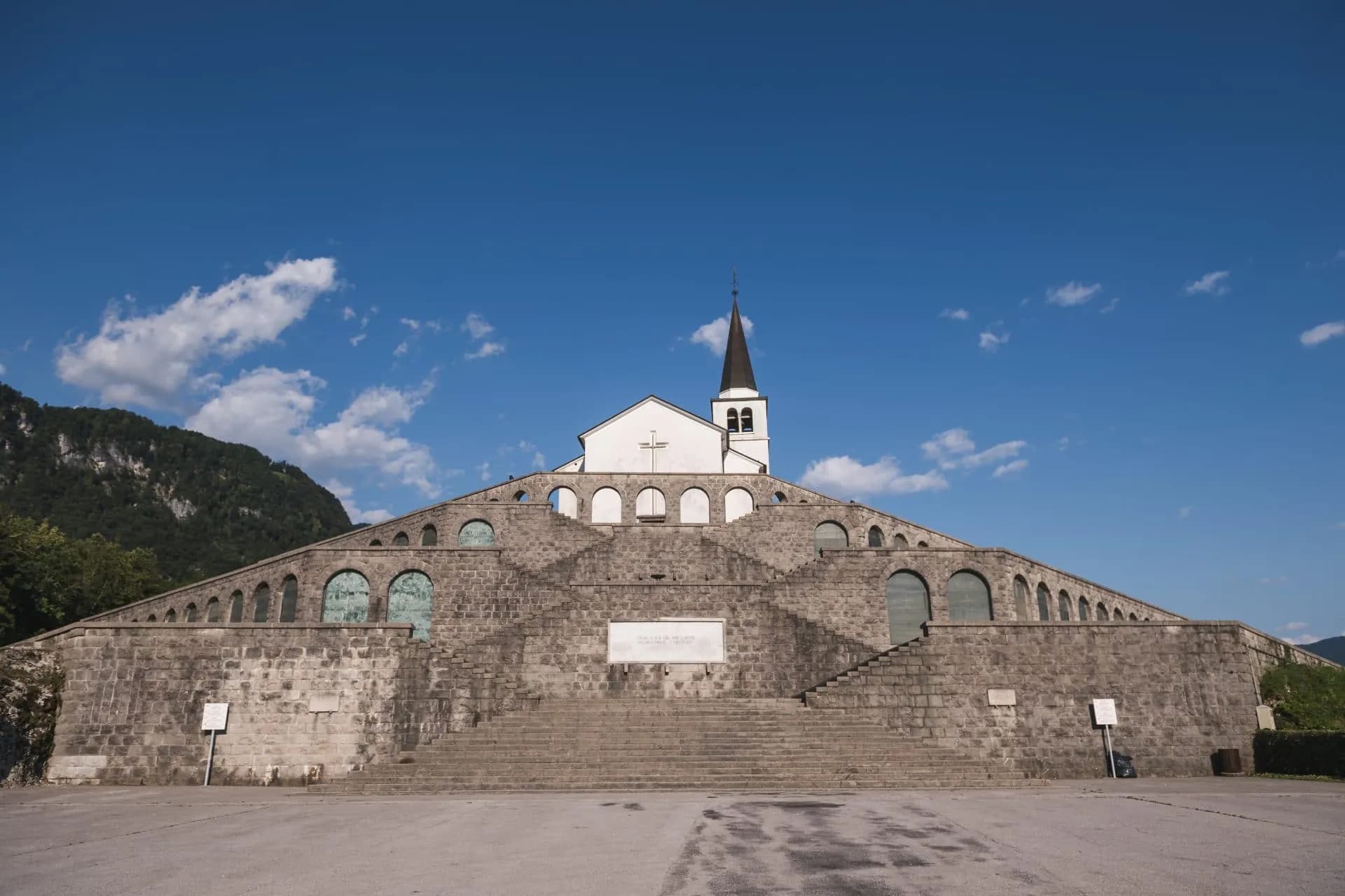 Kobarid Italian Ossuary WWI monument with tiered stone arches and white church against blue sky.