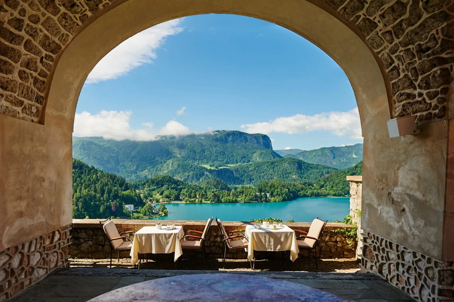 Outdoor dining tables framed by stone arch overlooking turquoise lake and green mountains, likely Lake Bled.