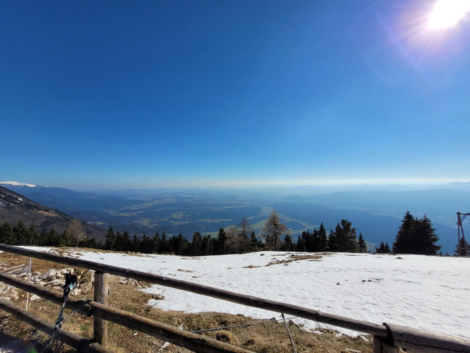 Mountain overlook with snow patches, wooden fence, and vast valley view under bright sun.