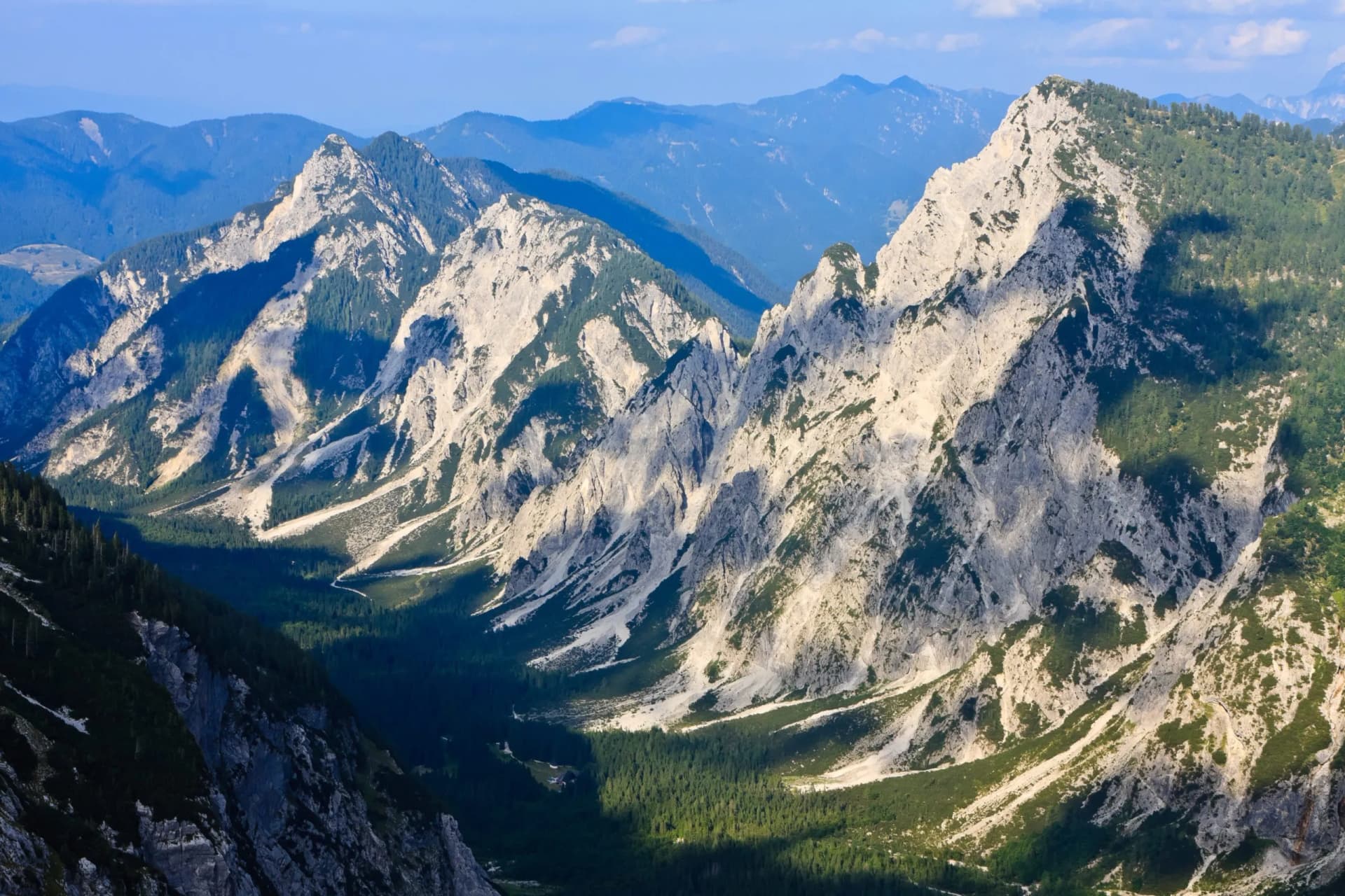 Rugged, sunlit limestone mountains with steep forested slopes and deep valleys under a blue sky.