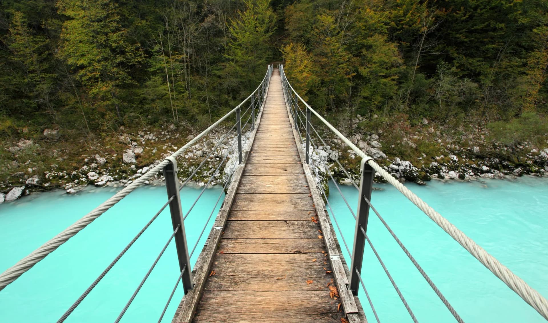 Wooden suspension bridge over bright turquoise river surrounded by dense forest.