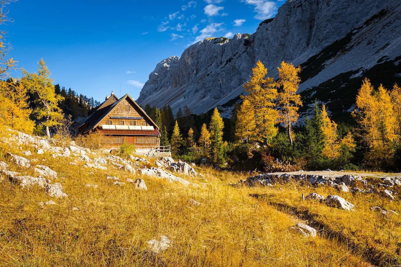Triglav Lakes Lodge in autumn with yellow trees and rocky mountains under blue sky.