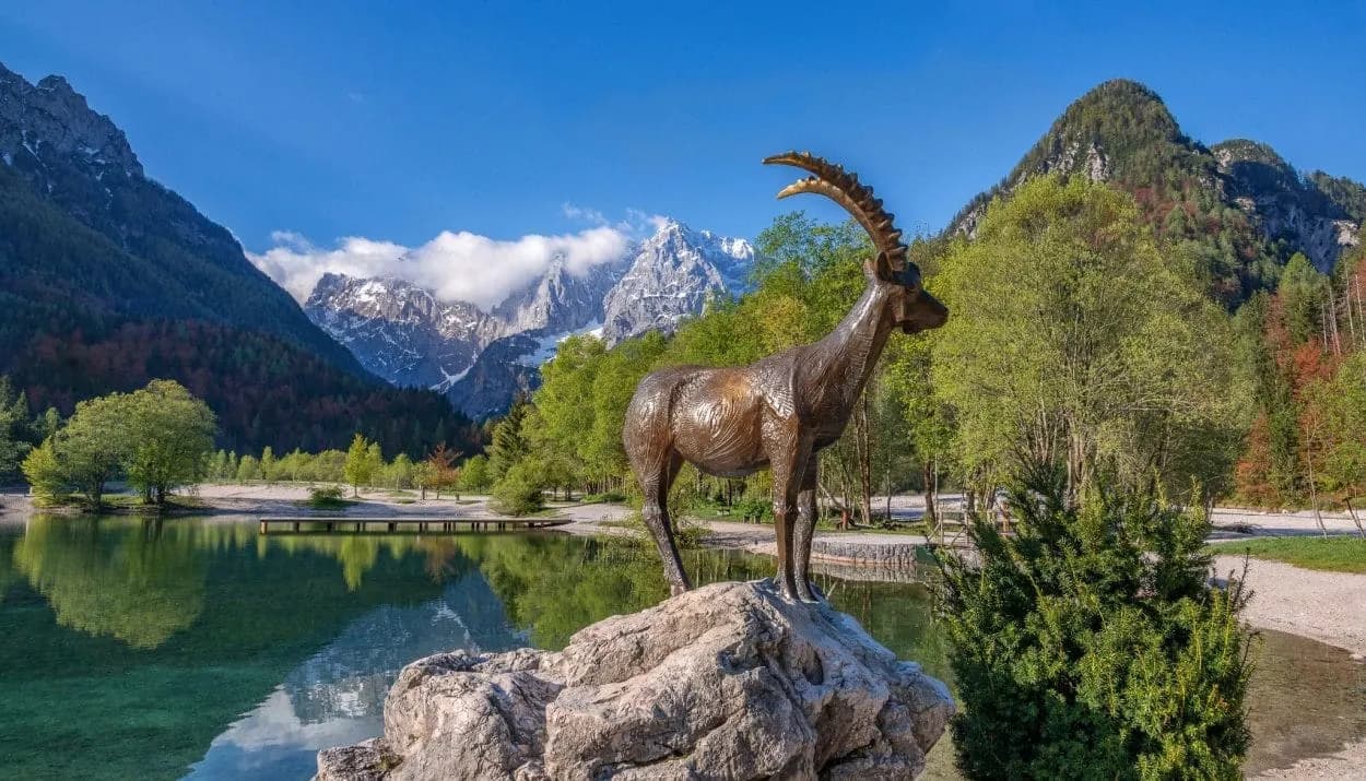 Bronze ibex statue by Lake Jasna near Kranjska Gora with snow-capped mountains.