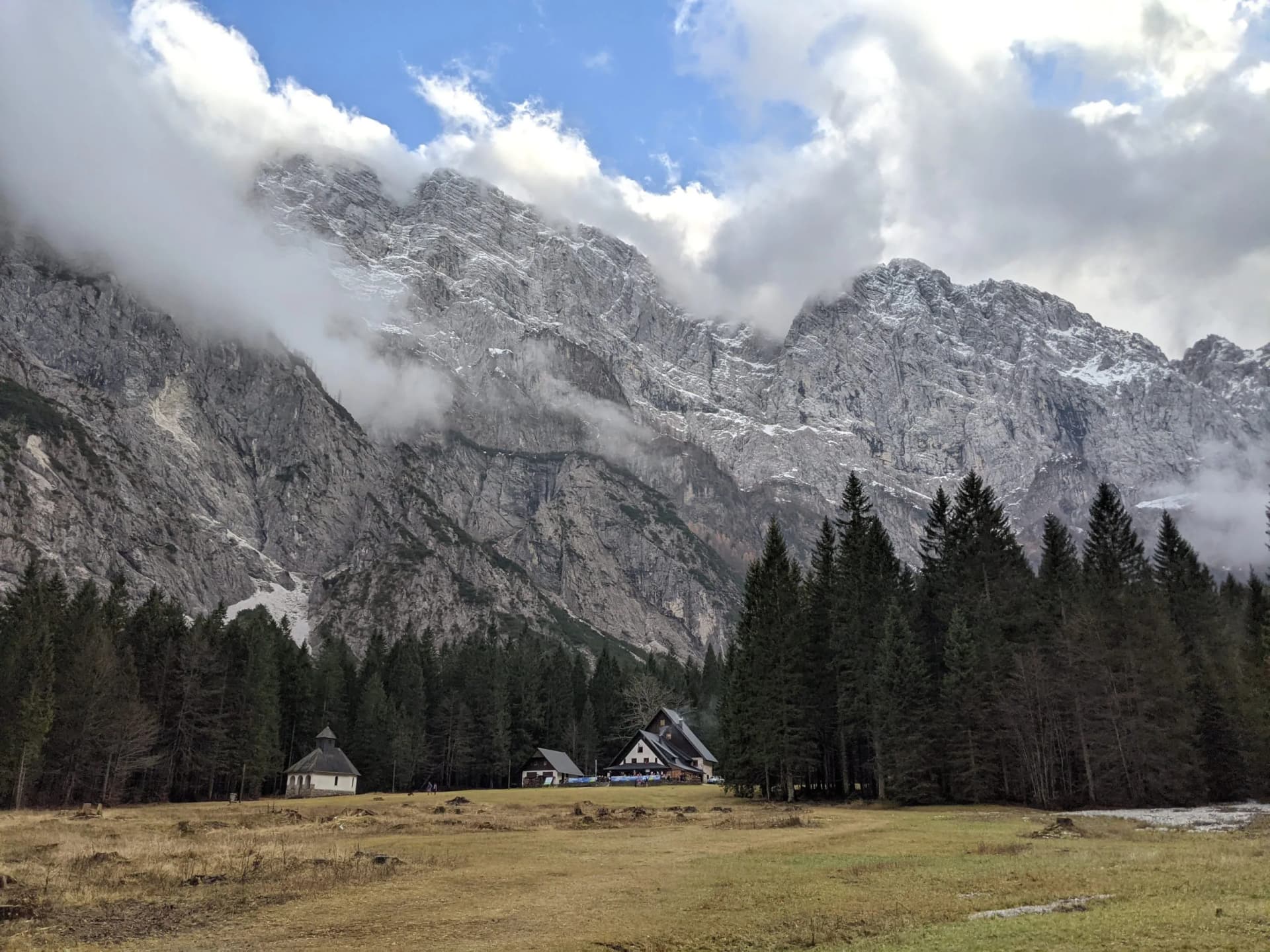 Alpine meadow with small buildings below steep, cloud-shrouded, snow-dusted mountains and pine forest.