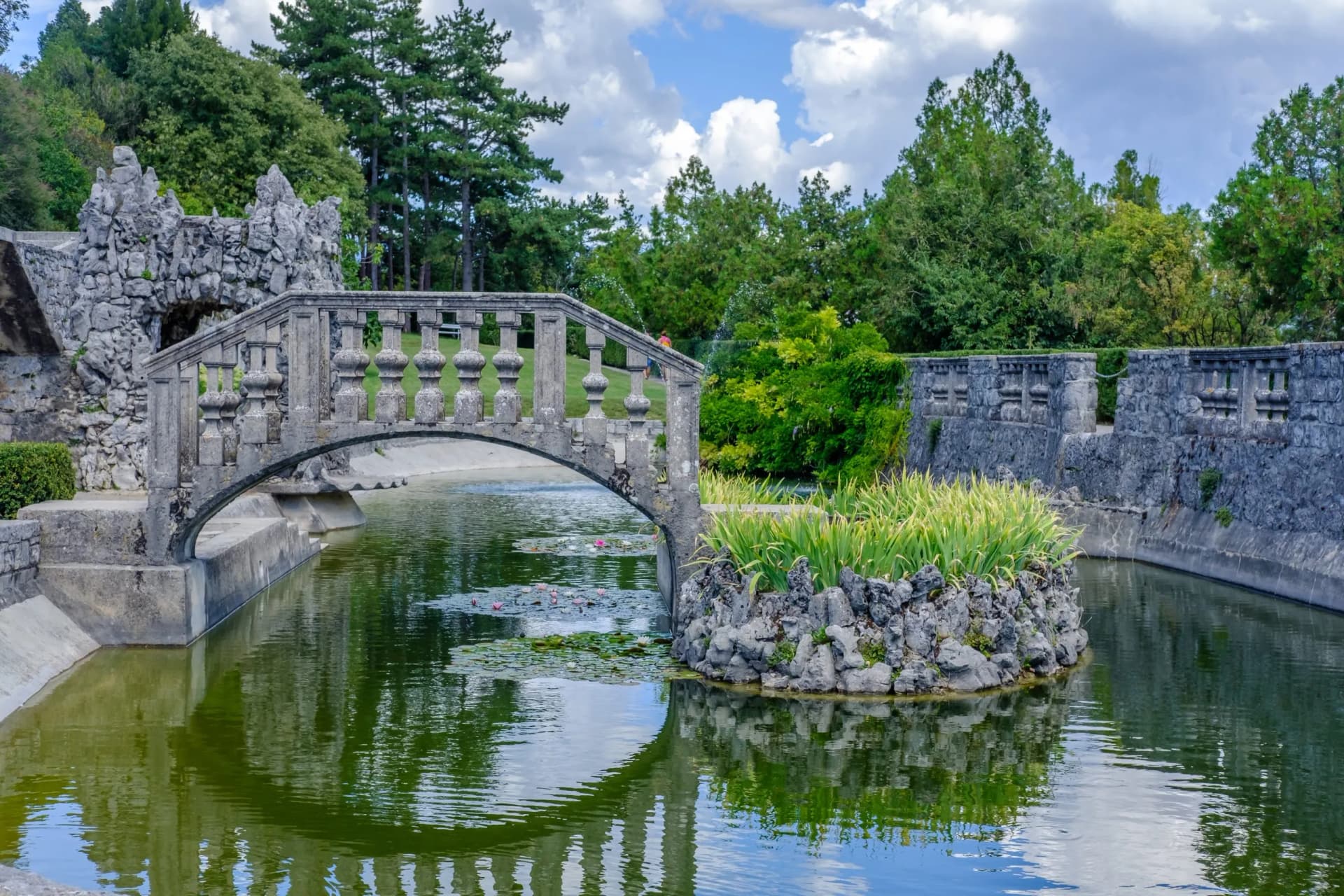 Stone bridge over canal with lily pads and lush green trees under cloudy sky.