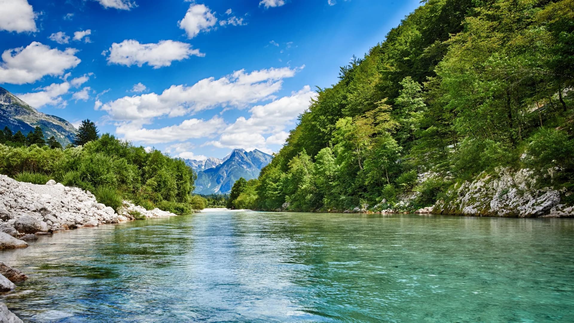 Clear river flowing between lush green banks toward distant, rugged mountains under a blue sky.