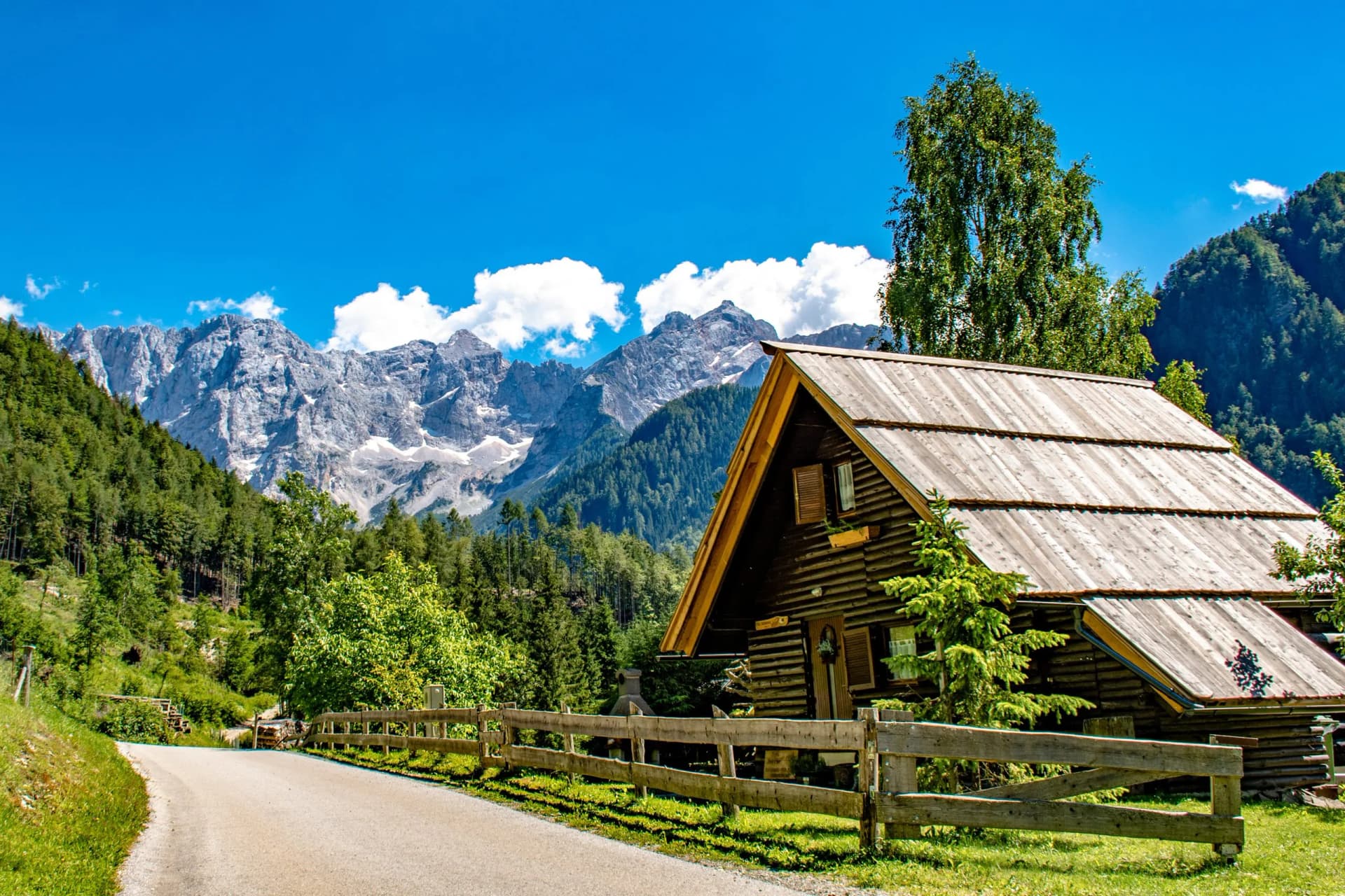 Quaint wooden alpine house near a road with snow-capped mountains under a blue sky in Zgornje Jezersko.