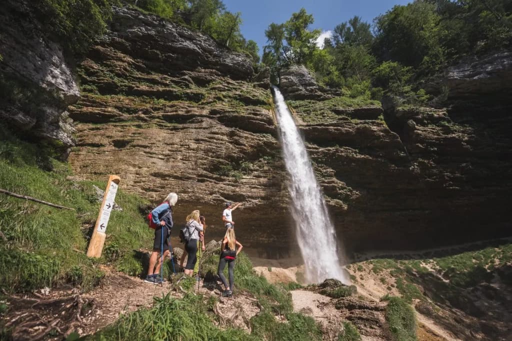 Hikers viewing Peričnik Waterfall cascading down a steep, layered rock face in a forest setting.