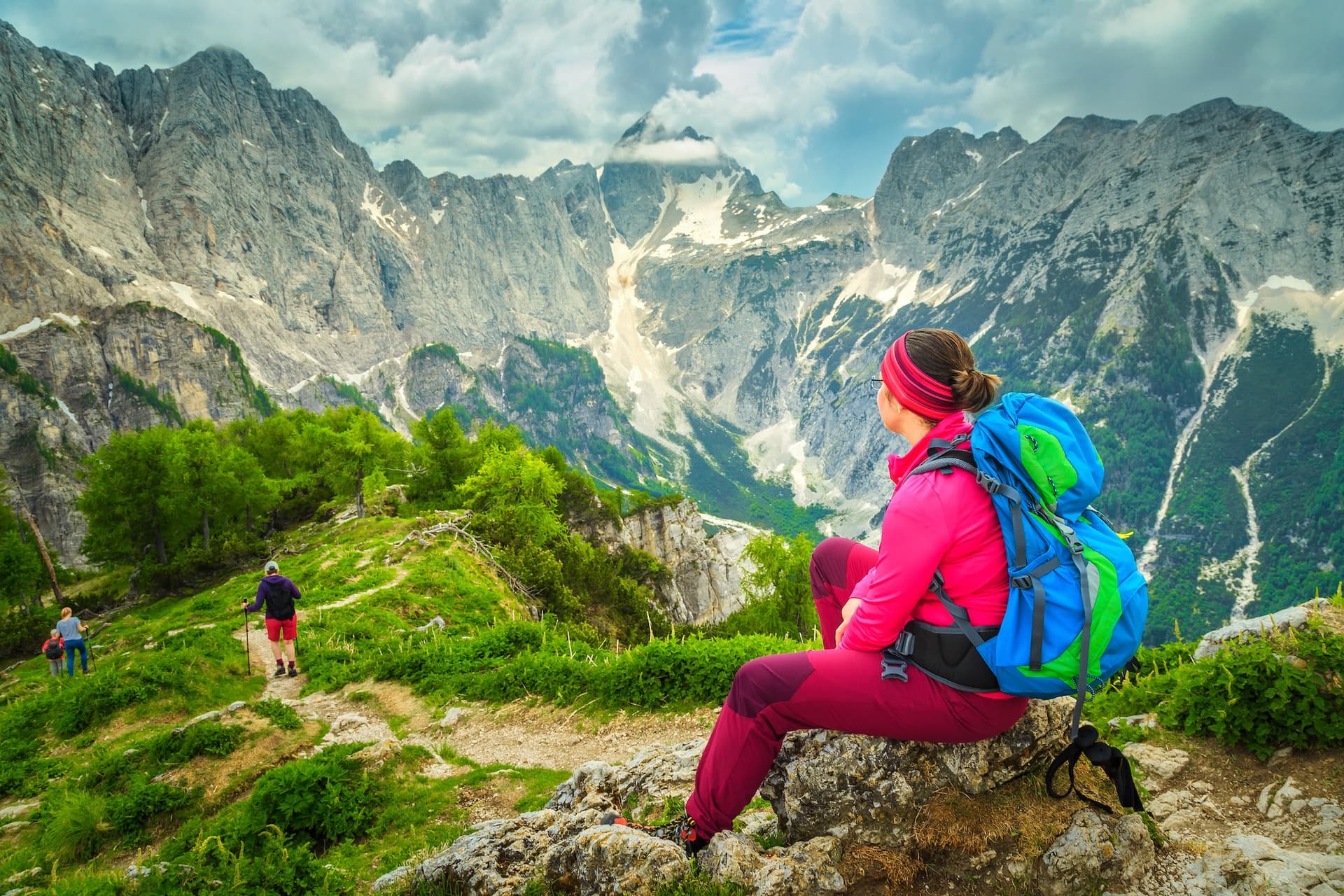 Hikers resting on a rocky outcrop overlooking steep, rocky mountains with patches of snow and green valleys.