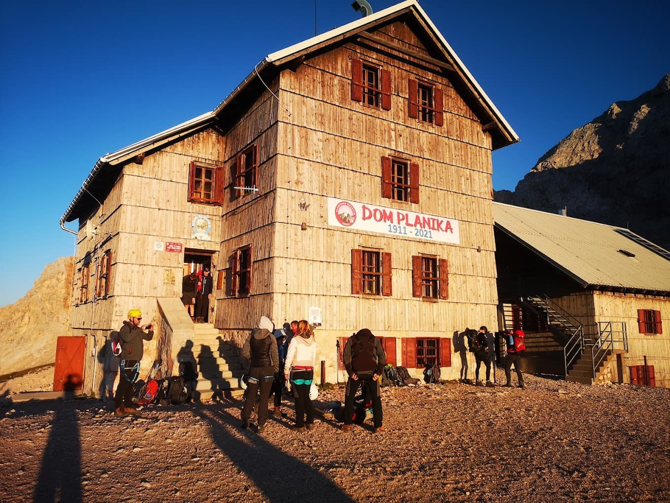 Dom Planika mountain hut with hikers gathering outside in morning sunlight near rocky peaks.